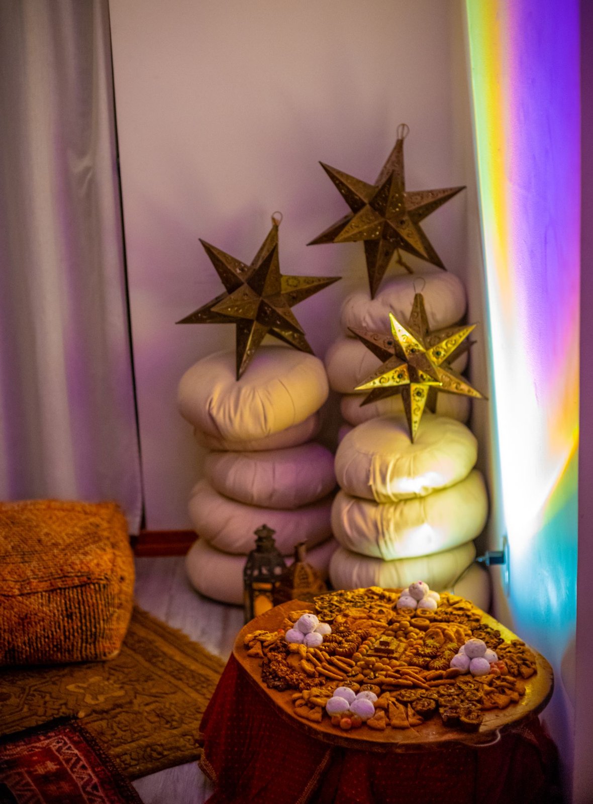 A stack of white cushions topped with metal star lanterns beside a platter of Moroccan sweets and a rainbow light
