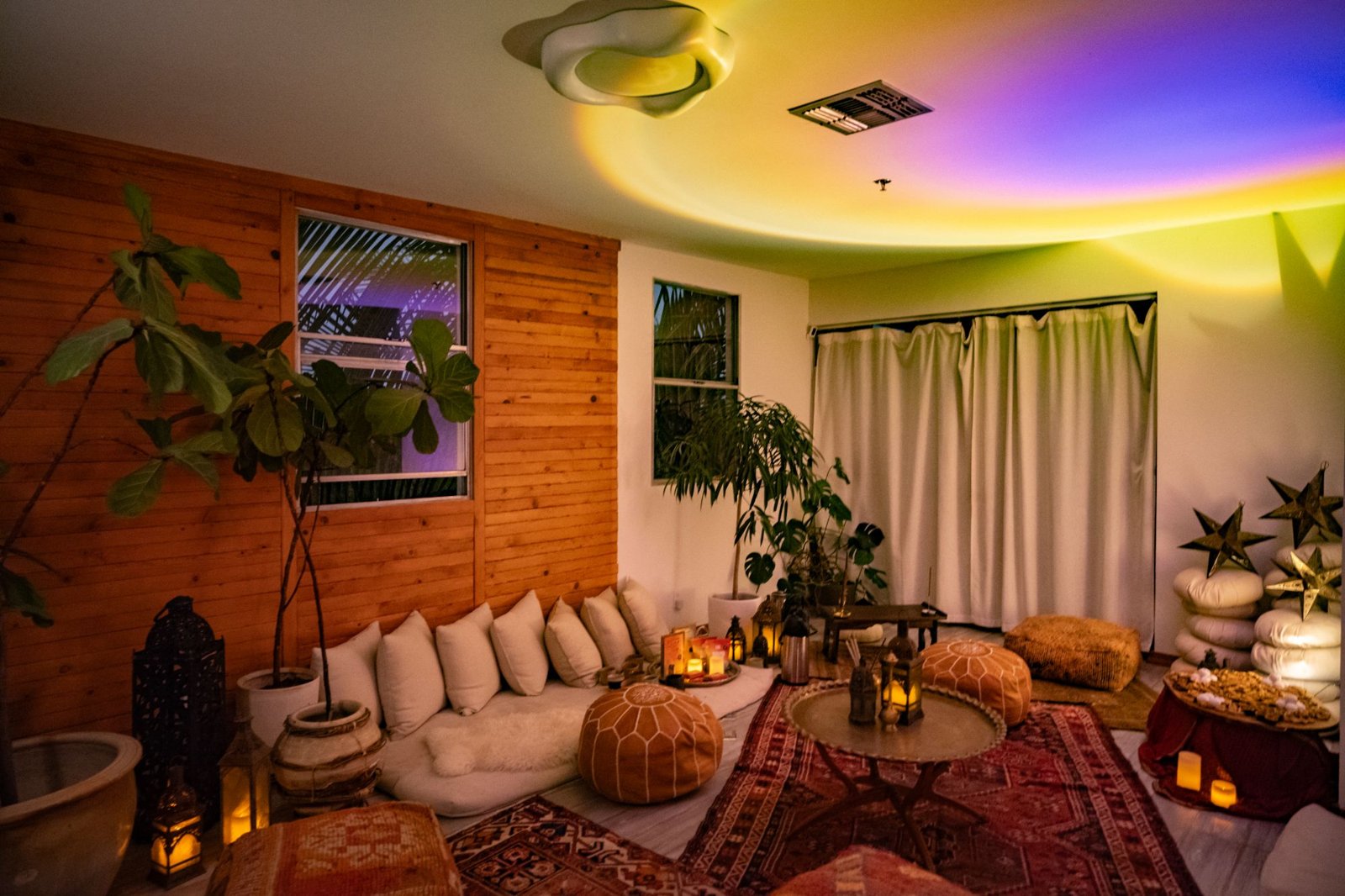 A lounge with white floor cushions, leather poufs, Persian rugs, lanterns, and a rainbow light across the ceiling
