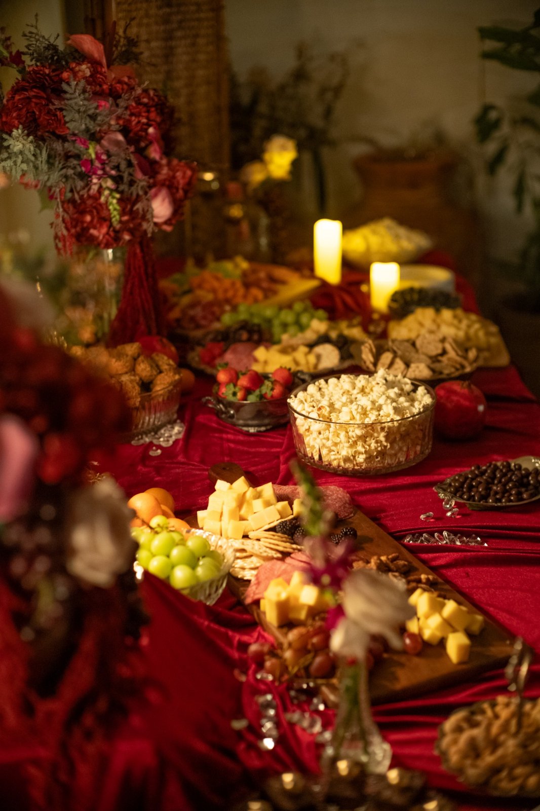 A grazing table on red velvet with cheeses, fruit, popcorn, sweets, candles, and red florals