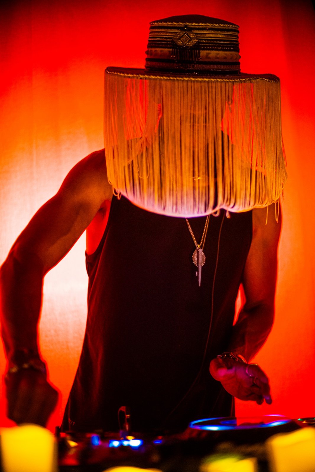 A DJ in a gold fringed hat at the decks against a red glowing curtain