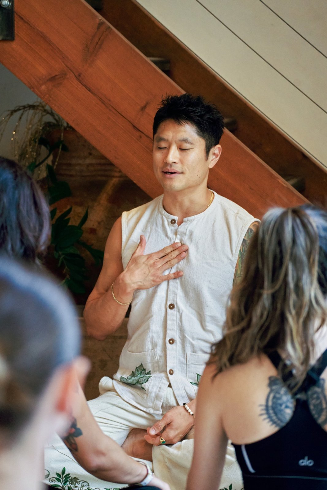 A man in a cream vest sitting cross-legged with his eyes closed and a hand on his chest, leading a meditation