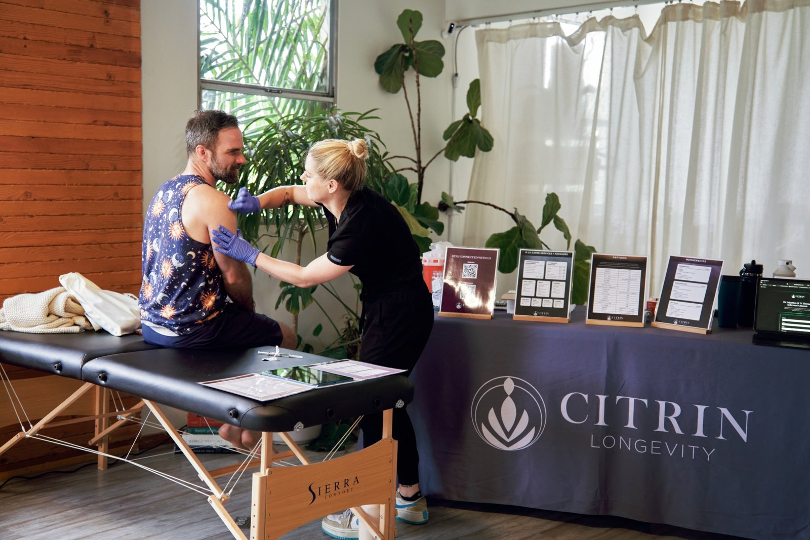 A nurse in gloves giving an injection to a seated man at the Citrin Longevity table, with menu cards on display
