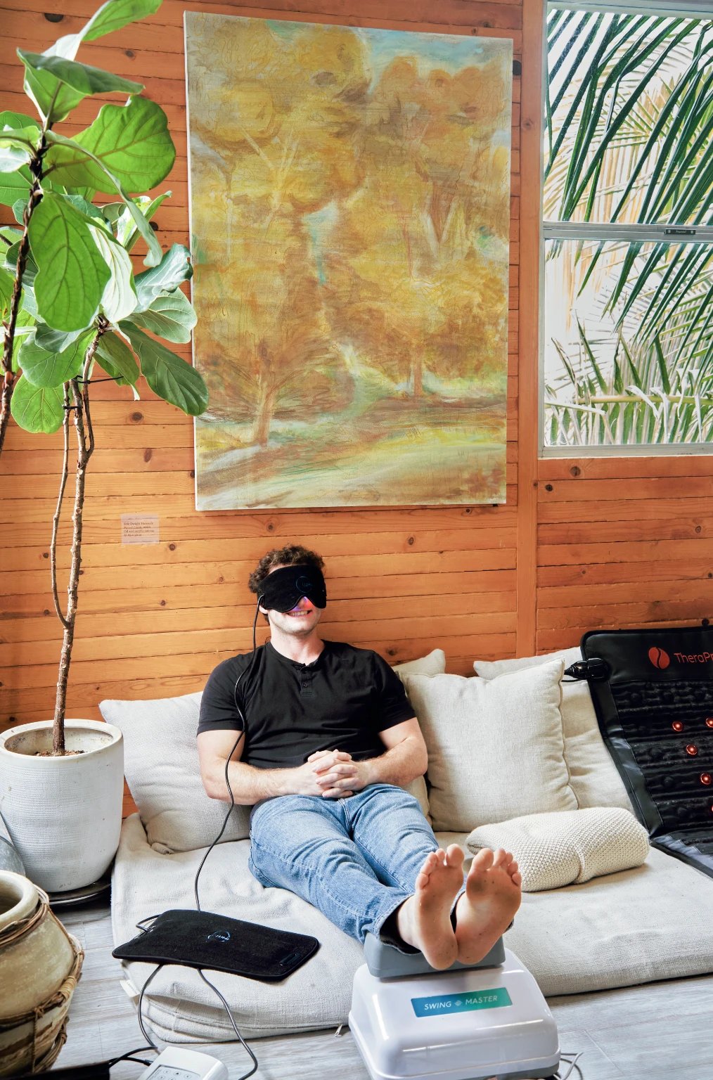 A man wearing a light-therapy eye mask and resting his feet on a massage machine on floor cushions beneath a painting
