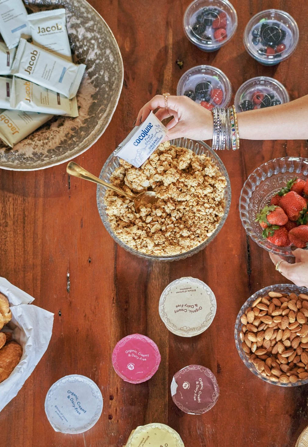 An overhead view of a breakfast table with granola, berries, almonds, yogurt cups, and protein bars