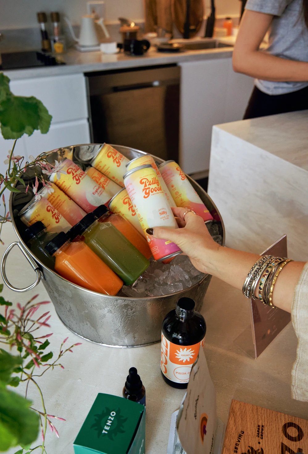 A hand reaching for a Psilly Goose can in an ice bucket of beverages and cold-pressed juices