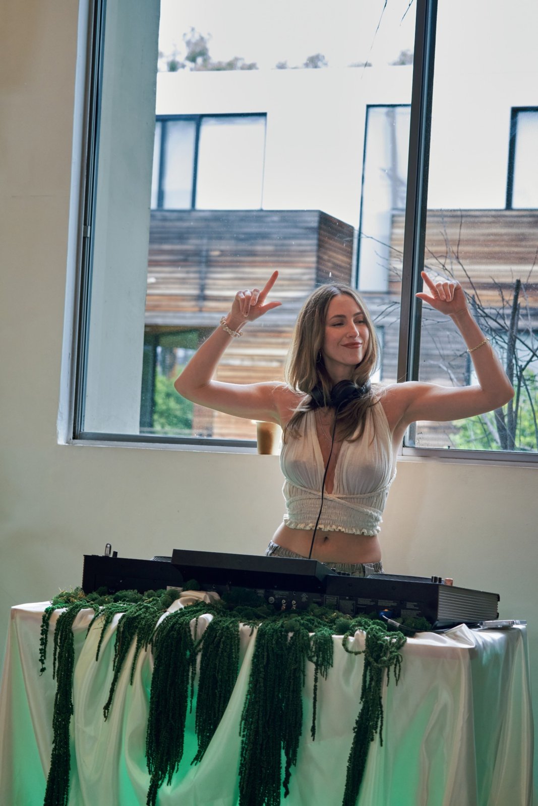 A DJ with headphones and raised pointing hands at turntables draped in white fabric and green garland