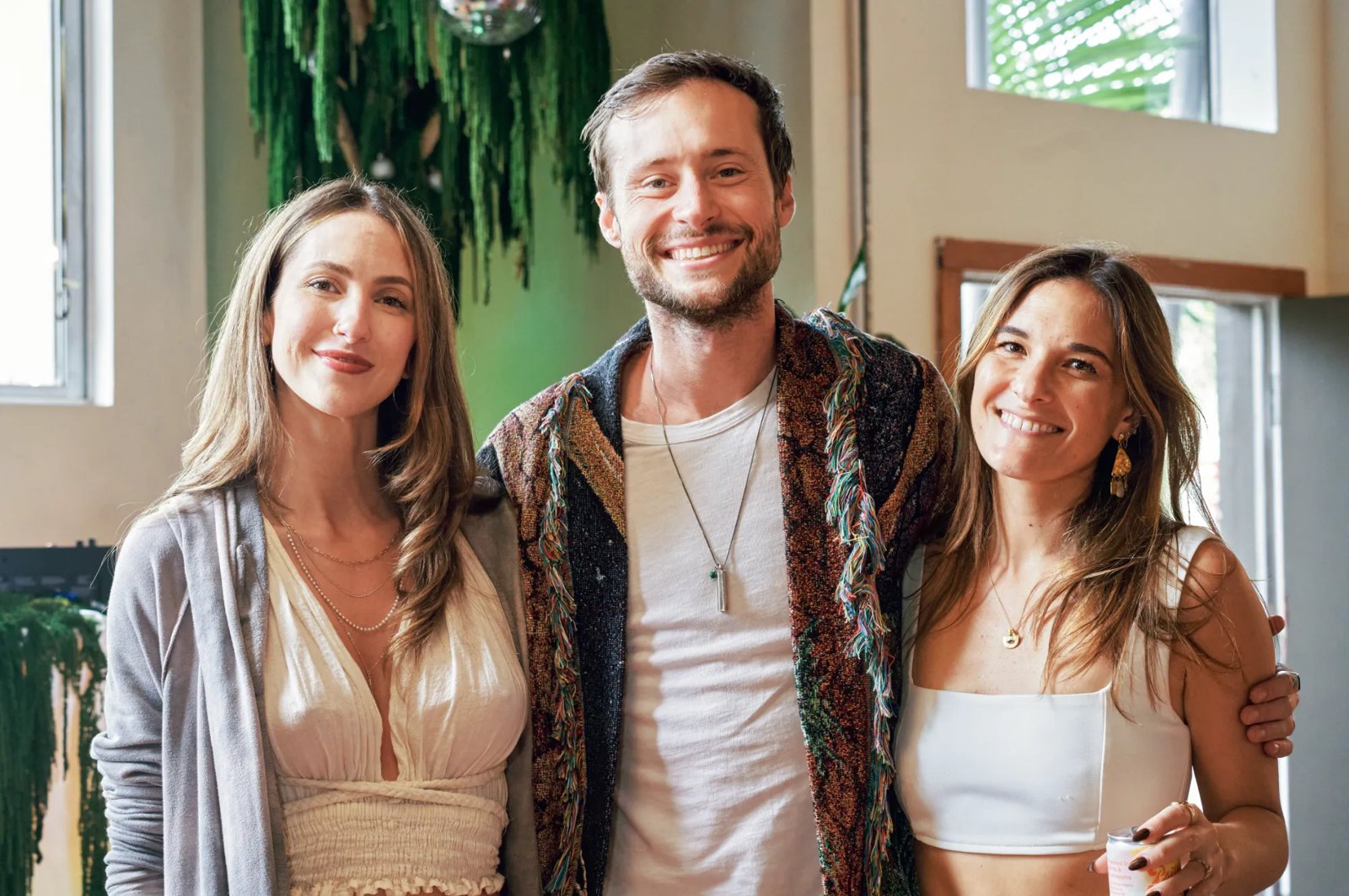Three smiling friends posing together in front of hanging green garland and a disco ball