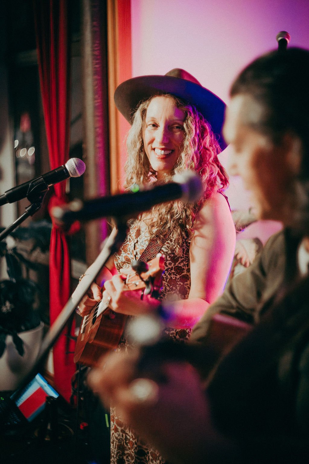 A singer in a wide-brim hat smiling as she plays a ukulele at a microphone, with red velvet curtains behind her