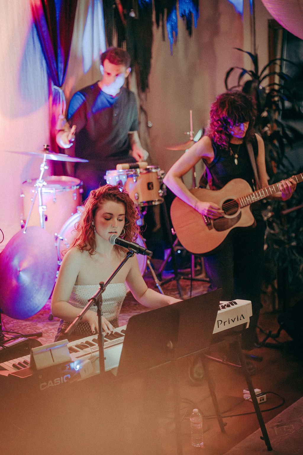 A band performing with a singer at a keyboard, a guitarist, and a drummer in front of draped fabric