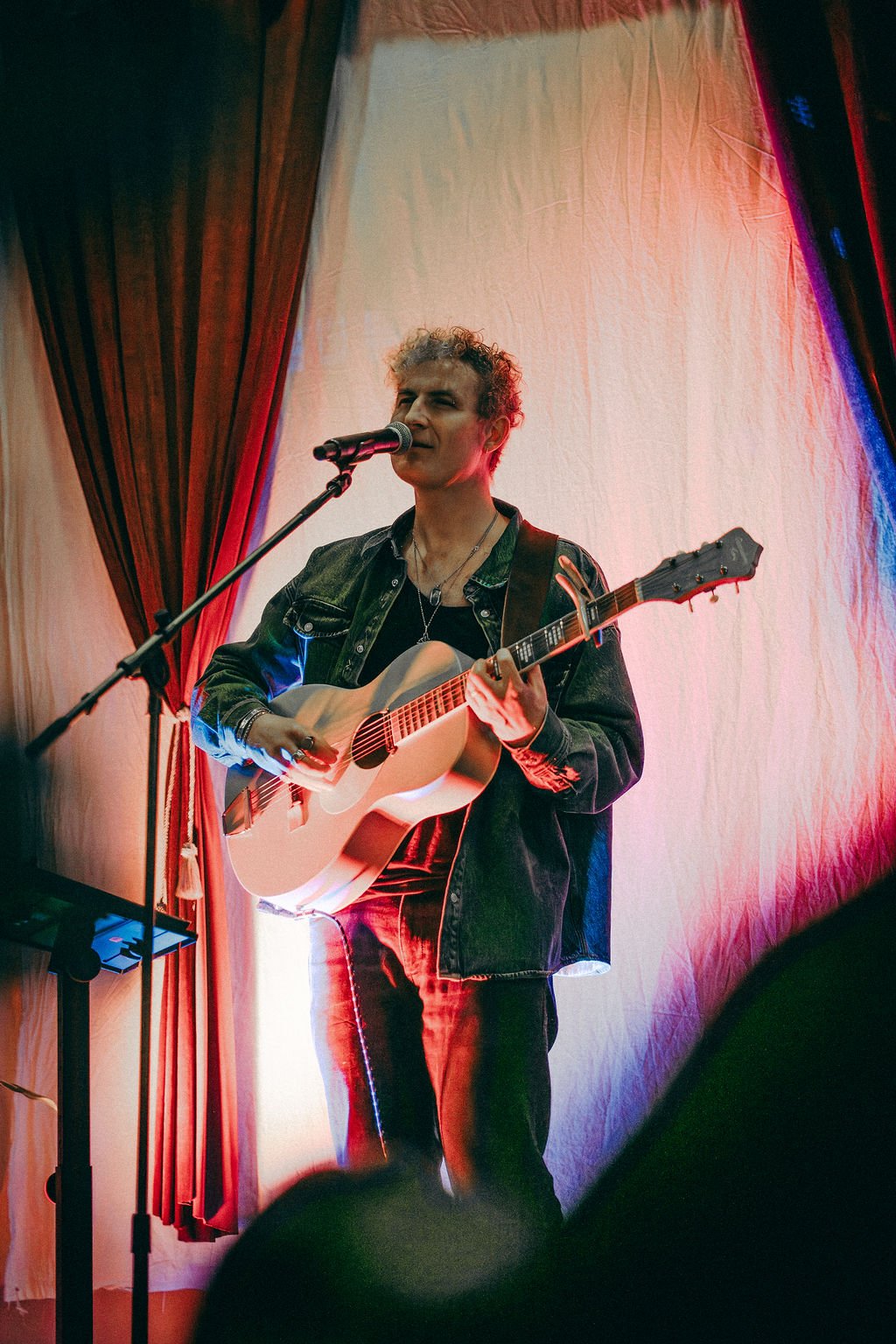 A singer-songwriter in a denim jacket playing an acoustic guitar at a microphone in front of red curtains and white fabric