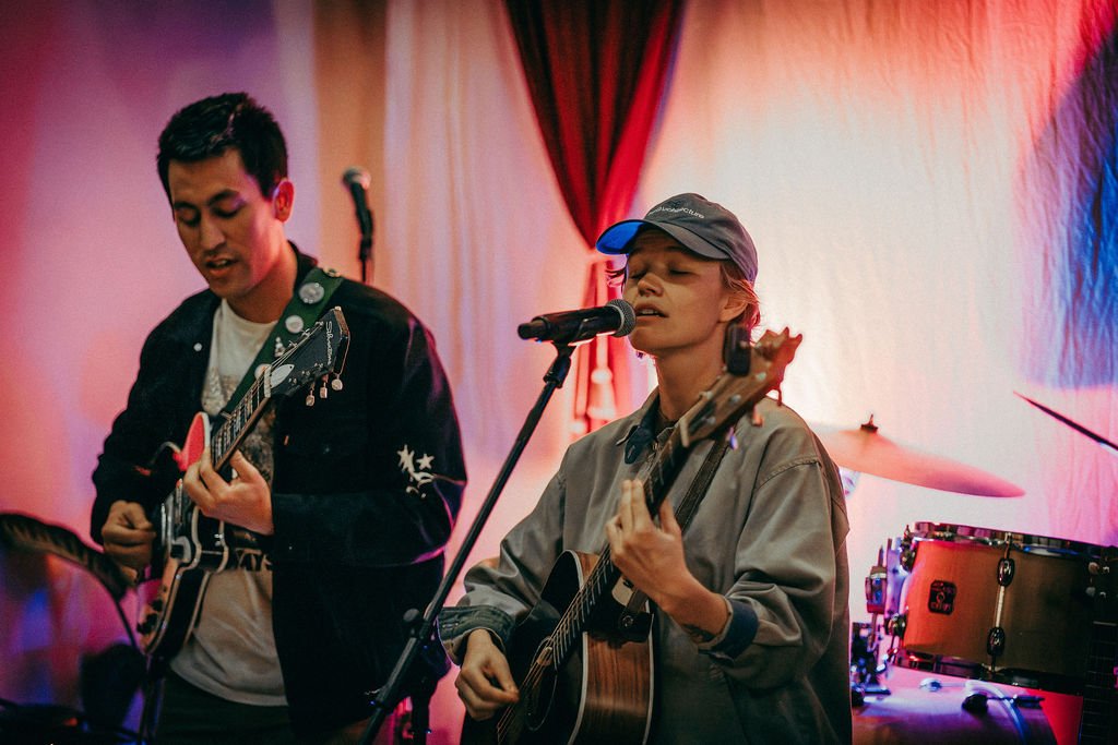 A woman in a striped jumpsuit singing into a microphone in front of the moss sign and a rack of sweatshirts