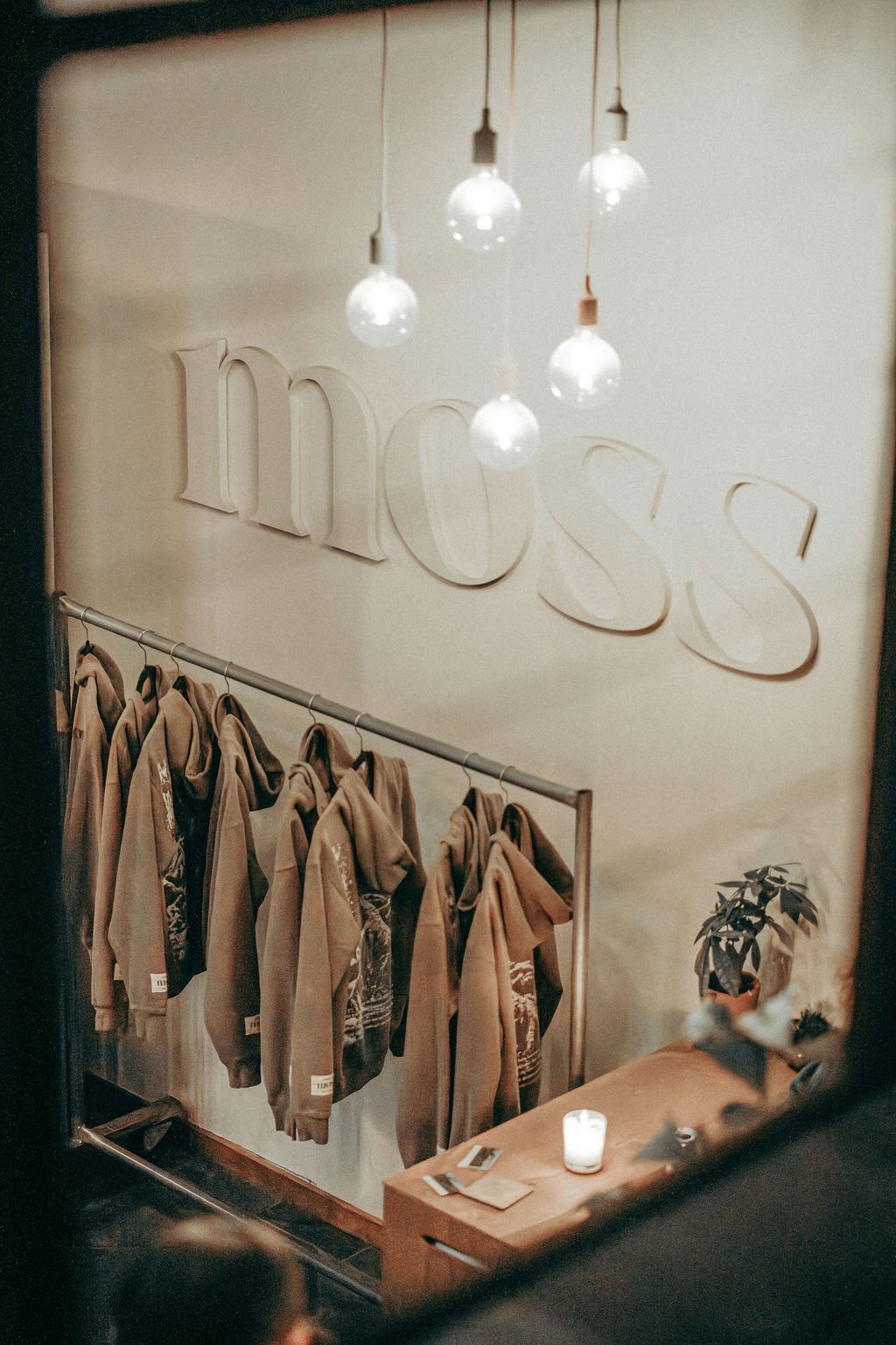 Hooded sweatshirts hanging on a rail beneath the Moss sign and a cluster of glass bulb pendants