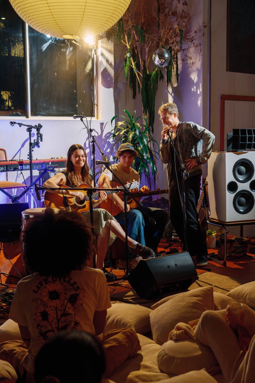 Two guitarists playing seated and a third performer singing into a microphone beside a large speaker, with a disco ball and green garland overhead and guests on cushions in the foreground
