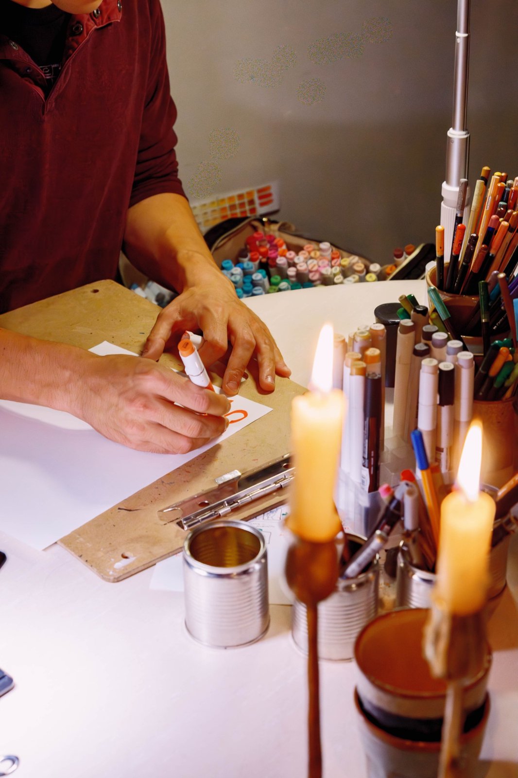 A live artist drawing on paper with a marker at a table of markers and pencils lit by two candles