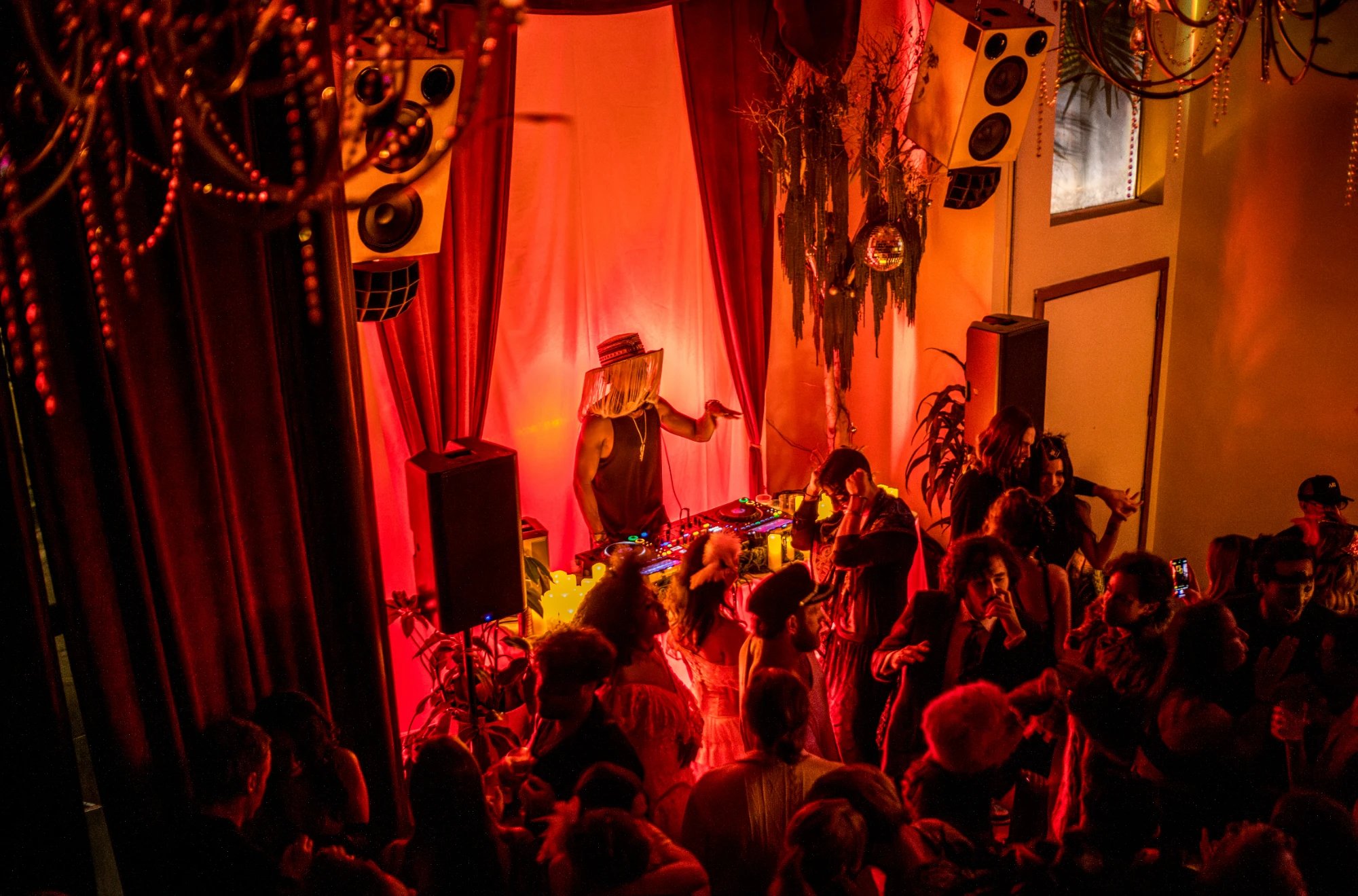 A DJ in a fringed hat playing for a dancing crowd in front of red velvet curtains, hanging speakers, a disco ball, and beaded chandeliers, seen from above