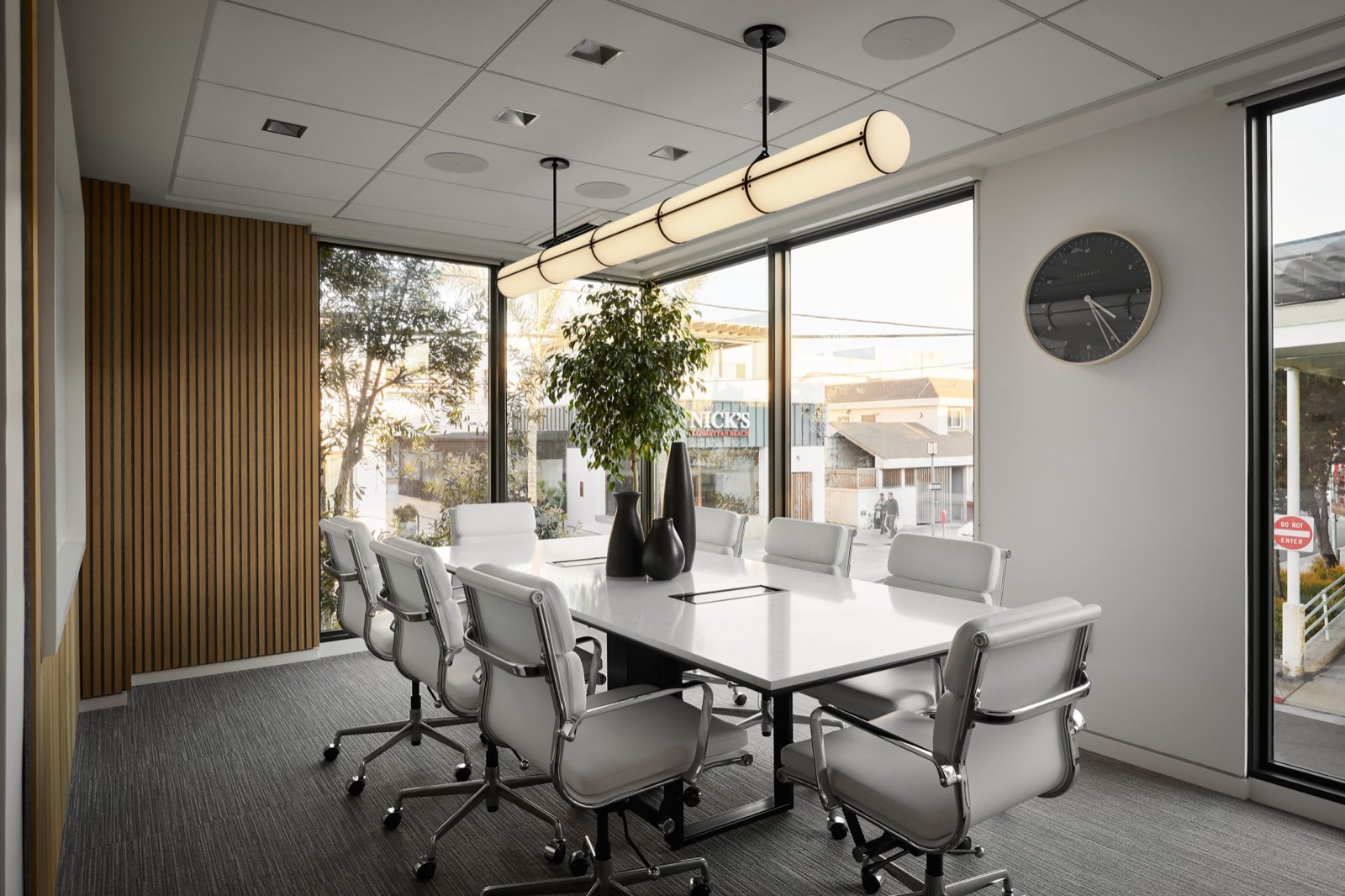 A conference room with a white stone table, white leather chairs, a long cylindrical pendant, and corner windows onto the street, beside a slatted wood wall