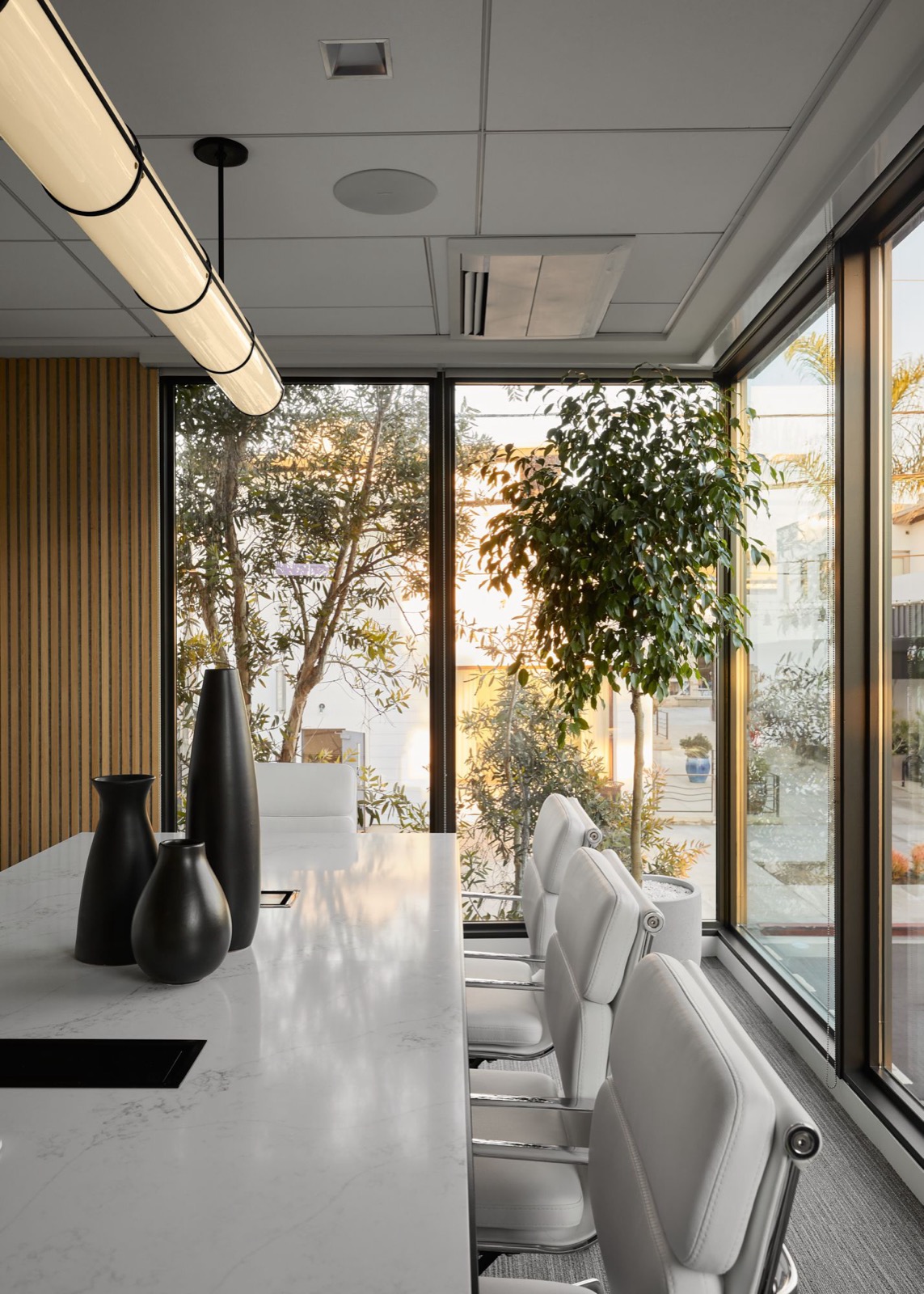 The head of the conference table with three black vases, white leather chairs, and corner windows with trees in the late light