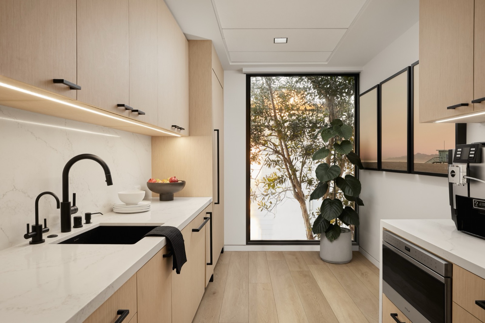 A galley kitchen with light oak cabinets, a white stone counter and backsplash, a black sink and faucets, and a tall window onto trees at sunset