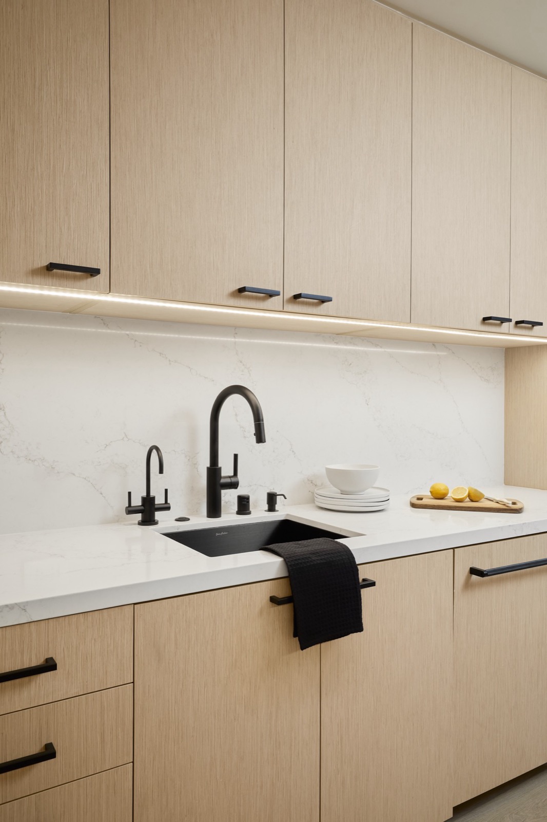 A kitchen wall of light oak cabinets with under-cabinet lighting, a marble backsplash, a black faucet, a stack of plates, and sliced lemons on a cutting board
