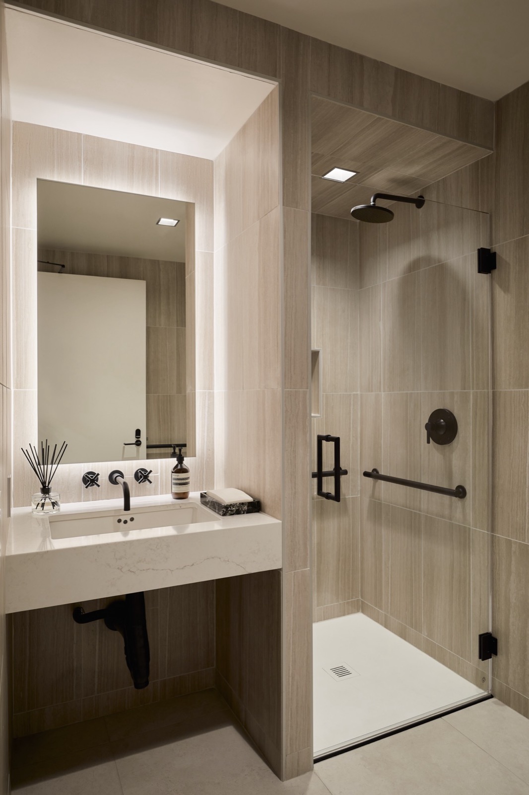 A bathroom with a backlit mirror over a floating stone vanity and a glass-door shower, both tiled in warm gray stone with black fixtures
