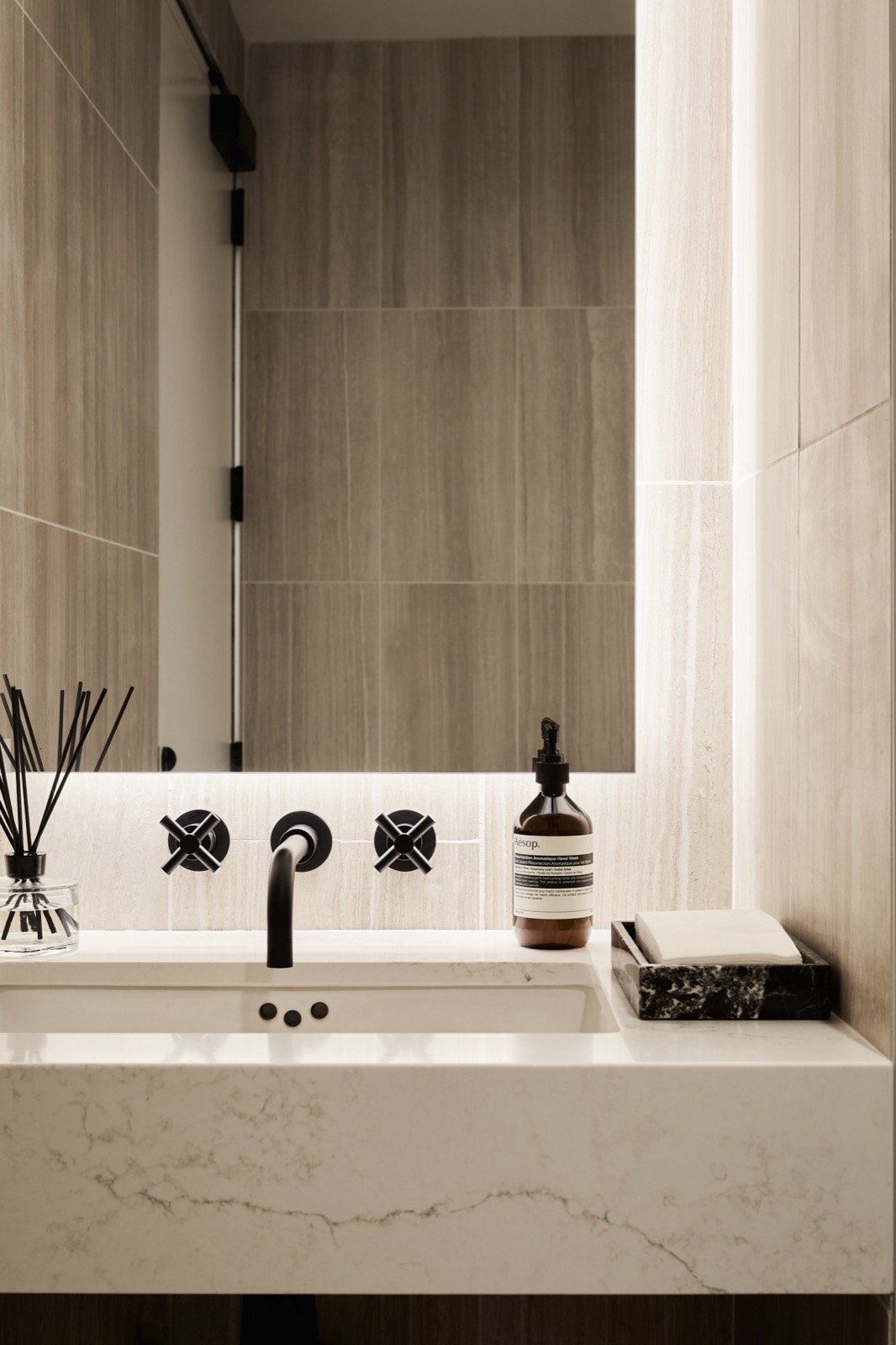 A detail of a wall-mounted black faucet with cross handles over a marble-look vanity, with a backlit mirror and a hand wash bottle