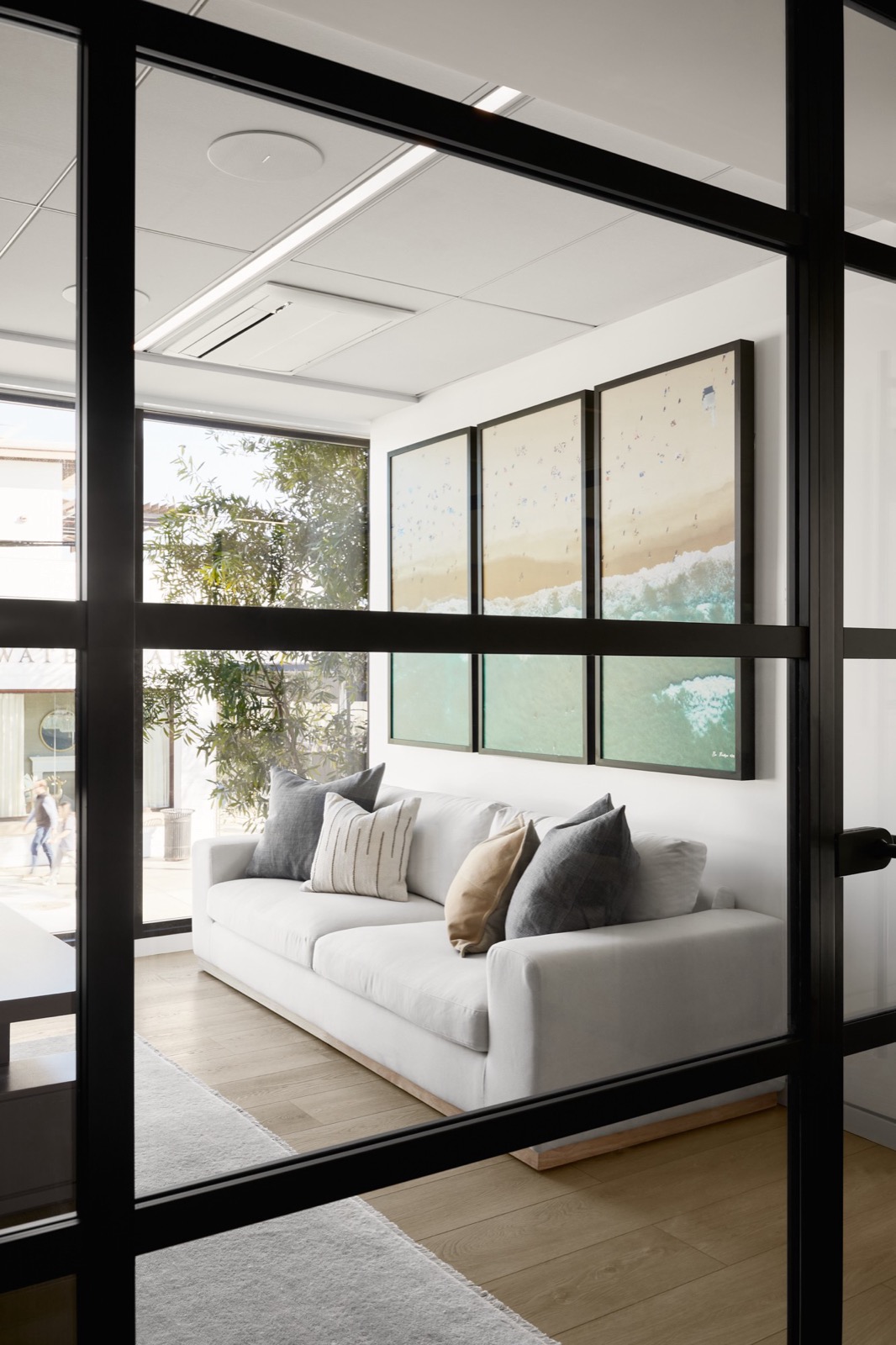 A small lounge seen through a black-framed glass wall, with a white sofa and a triptych aerial beach print above it, beside a corner window onto trees