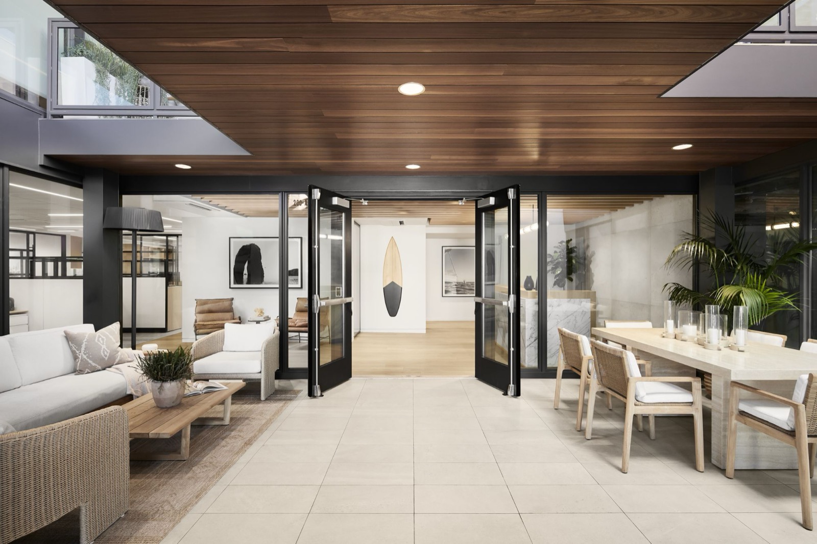 The open double doors of the office entry beneath a wood plank ceiling, with a surfboard on the wall inside, a patio lounge to the left, and a long travertine dining table with candle lanterns to the right