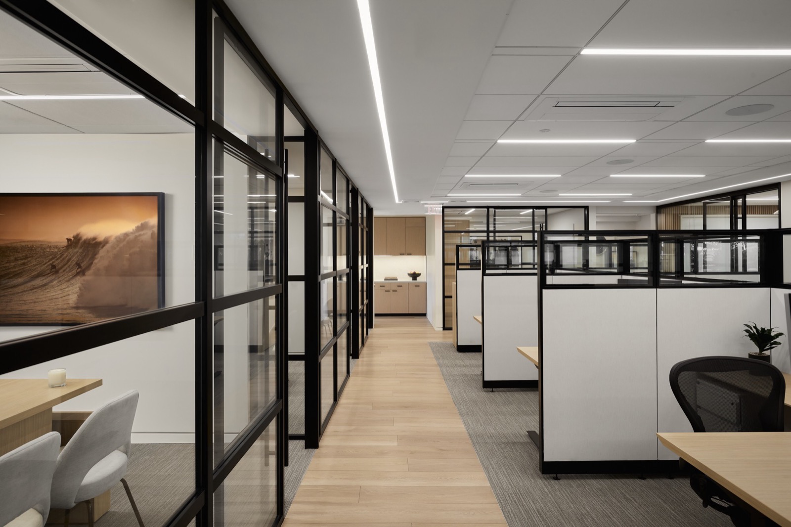 A corridor between black-framed glass offices and open workstations, with a framed print of a surfer on a large wave on the left and a kitchenette at the far end