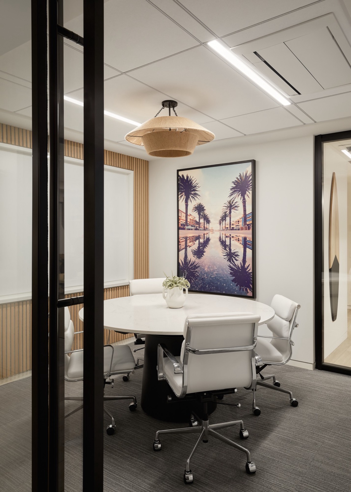 A small meeting room seen through a black-framed glass door, with a round white table, white leather chairs, a woven pendant, and a framed print of palm trees reflected in water
