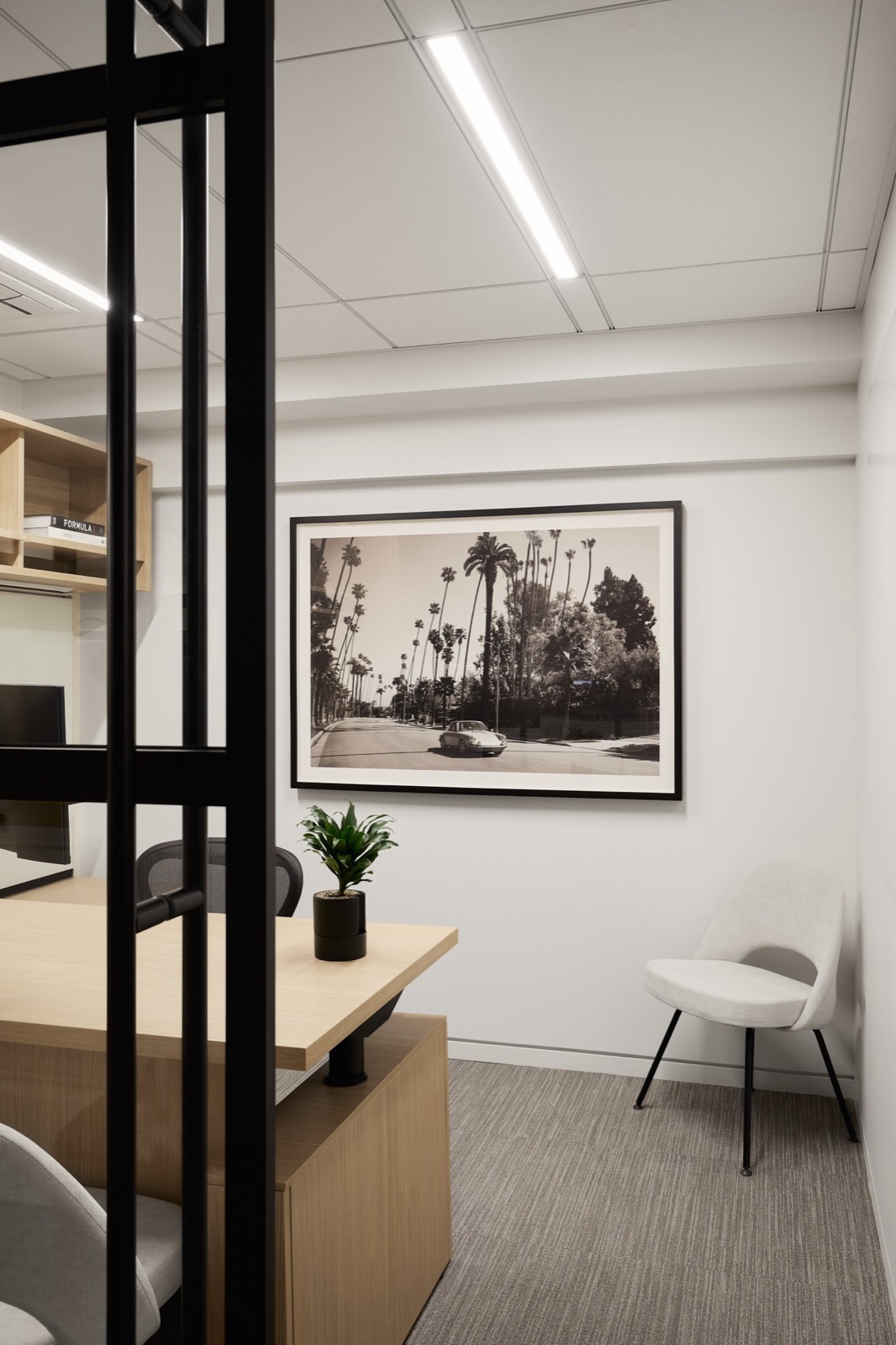 A private office seen through a black-framed glass door, with a light wood desk, a gray chair, a green plant, and a framed black-and-white print of a car on a palm-lined street