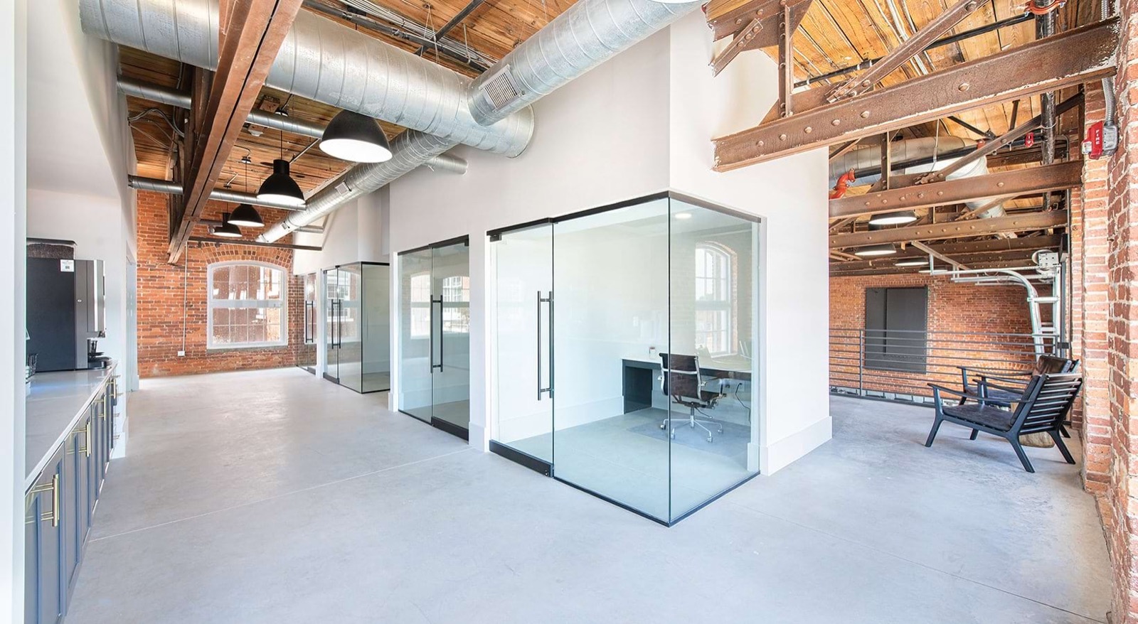 A corridor of glass-walled offices beneath exposed timber, steel beams, and ductwork, with a coffee station and brick walls