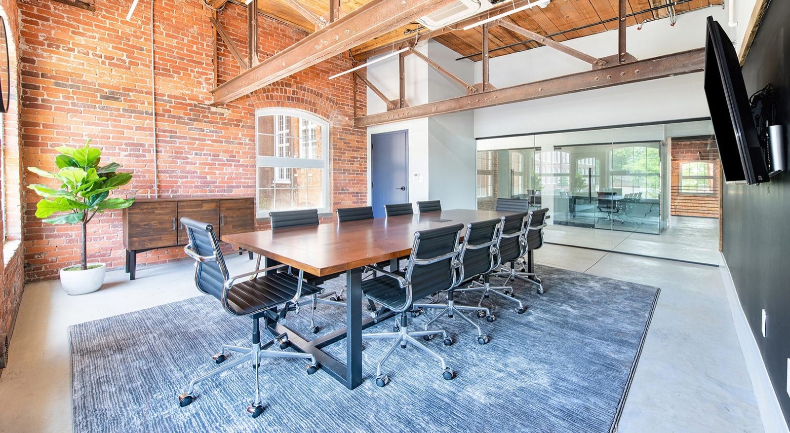 A conference room with a long wood table, black leather chairs, a brick wall, and a glass-walled meeting room beyond