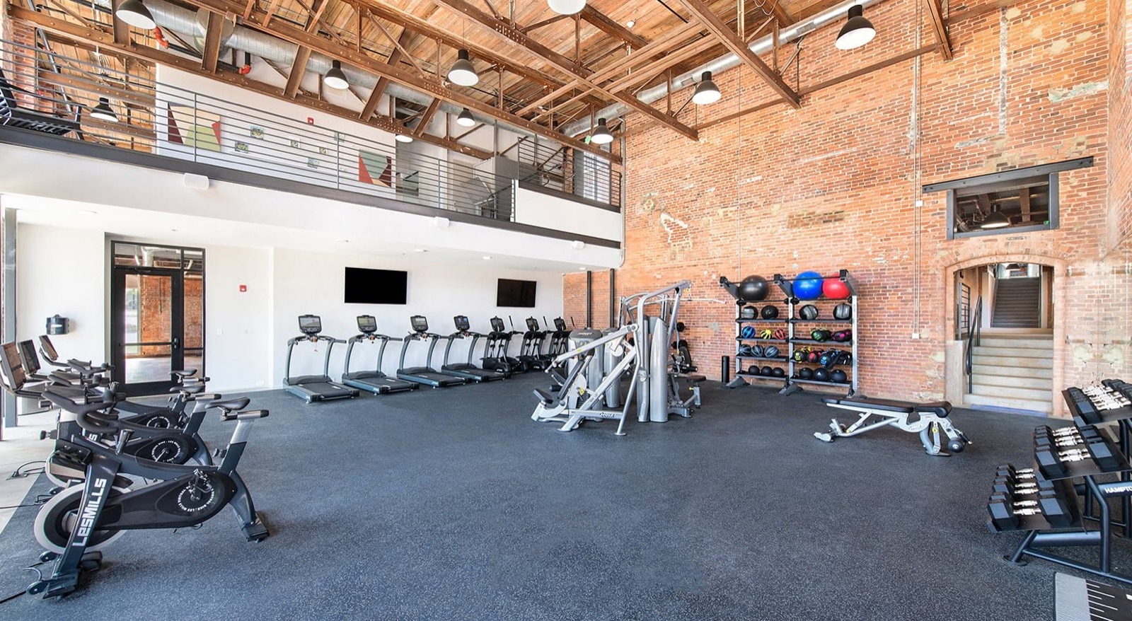 A double-height fitness center with spin bikes, treadmills, and a medicine-ball rack beside a brick wall and a mezzanine