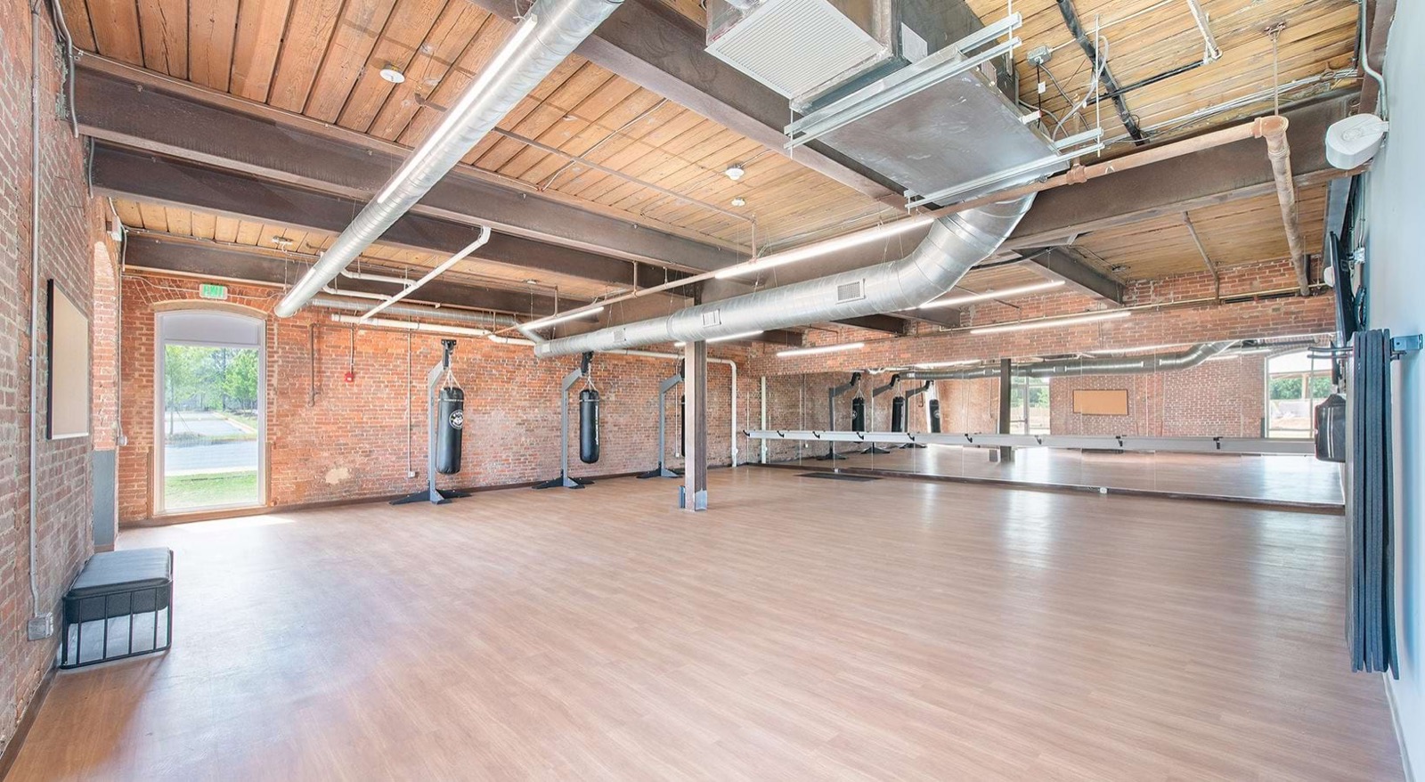 A studio with a wood floor, hanging heavy bags, a mirrored wall with a barre, and exposed timber and ducts
