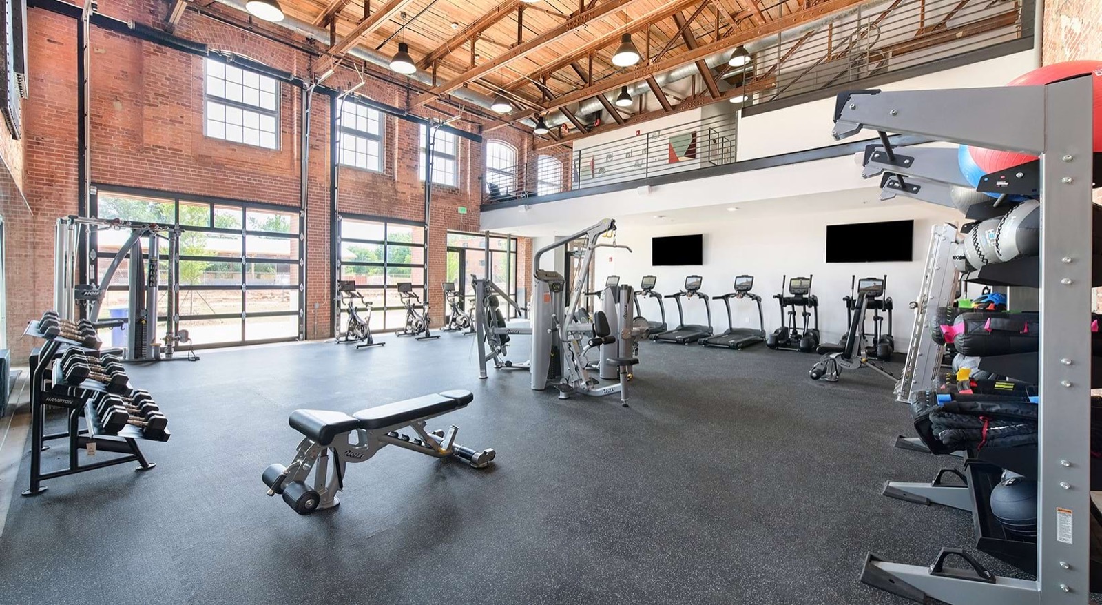 A fitness center with dumbbell racks, strength machines, and spin bikes in front of glass garage doors and brick walls