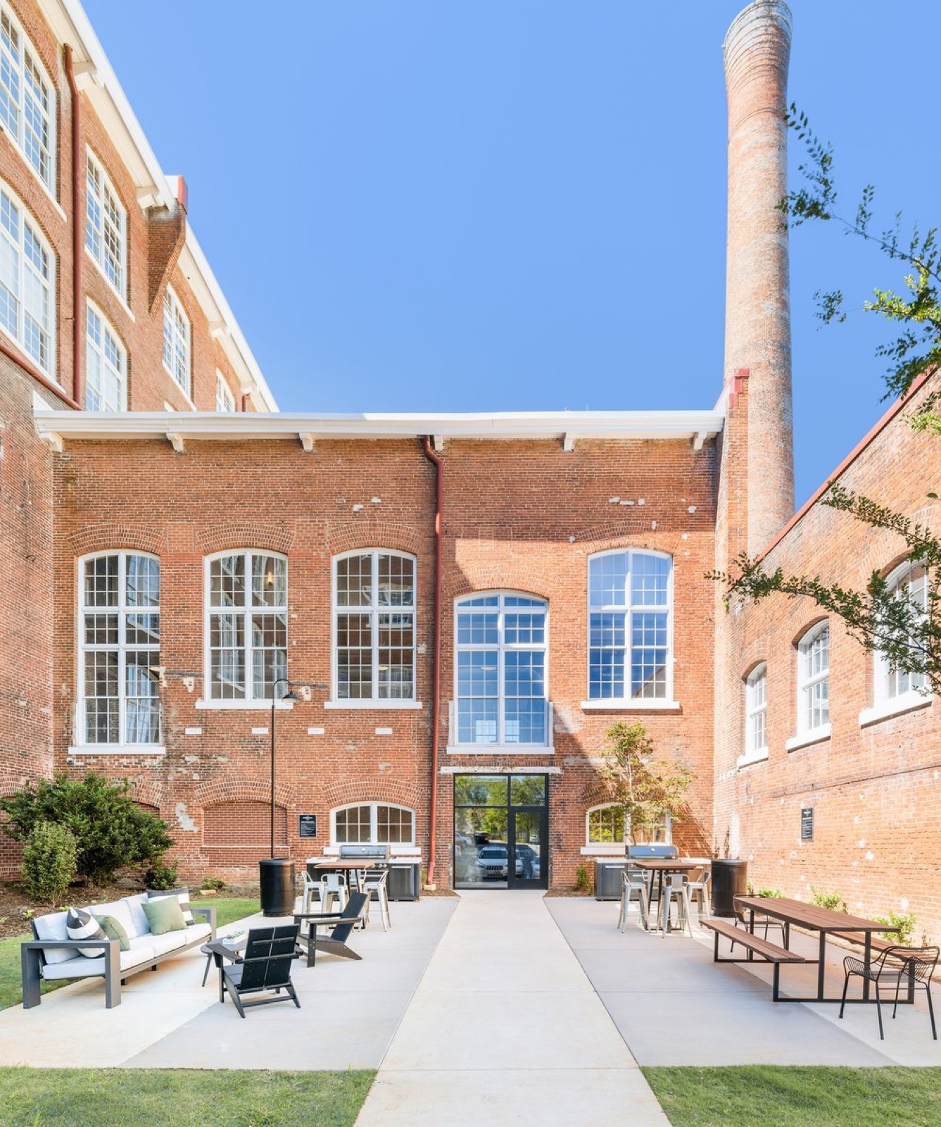 A courtyard with lounge seating, bar tables, and grills in front of the mill and its smokestack