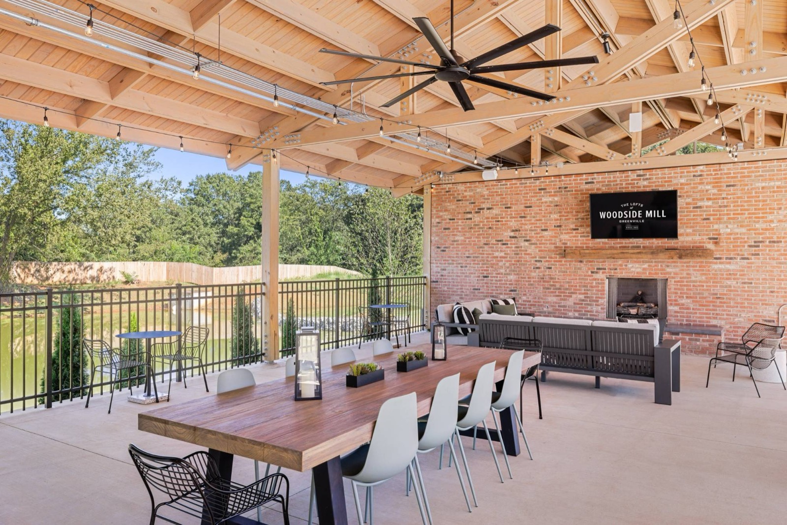 A covered pavilion with a long wood dining table, a sofa in front of a brick fireplace, and a view of the pond