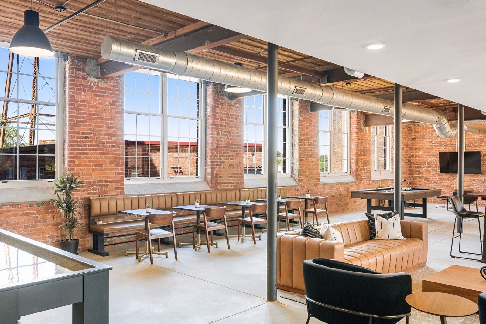 Brick-walled lounge with a long leather banquette, café tables, and a pool table beneath exposed wood beams and ductwork