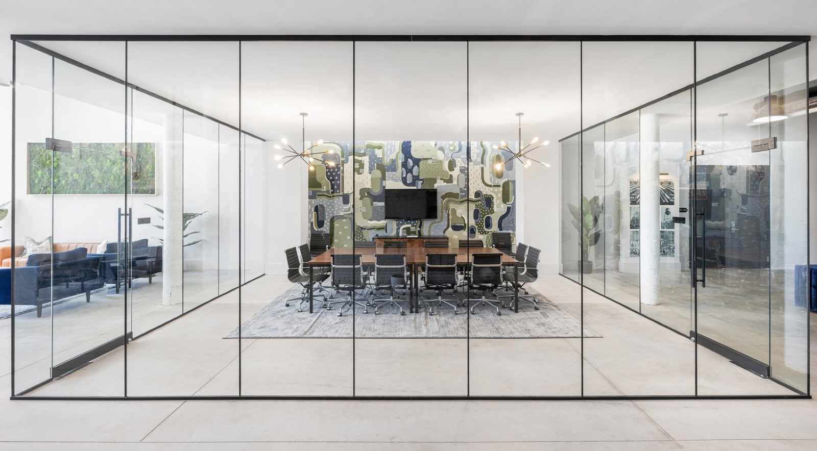 A glass-walled conference room with a long wooden table beneath two sputnik chandeliers and a patterned mural wall