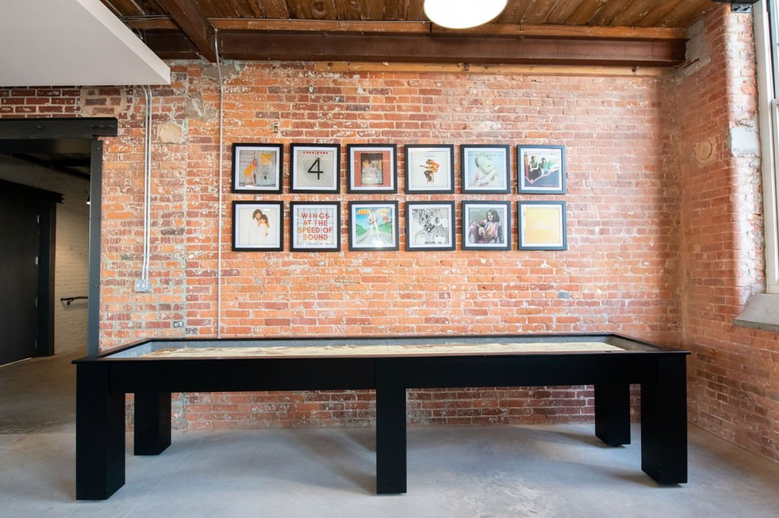 A black shuffleboard table in front of a brick wall hung with a grid of framed vinyl album covers
