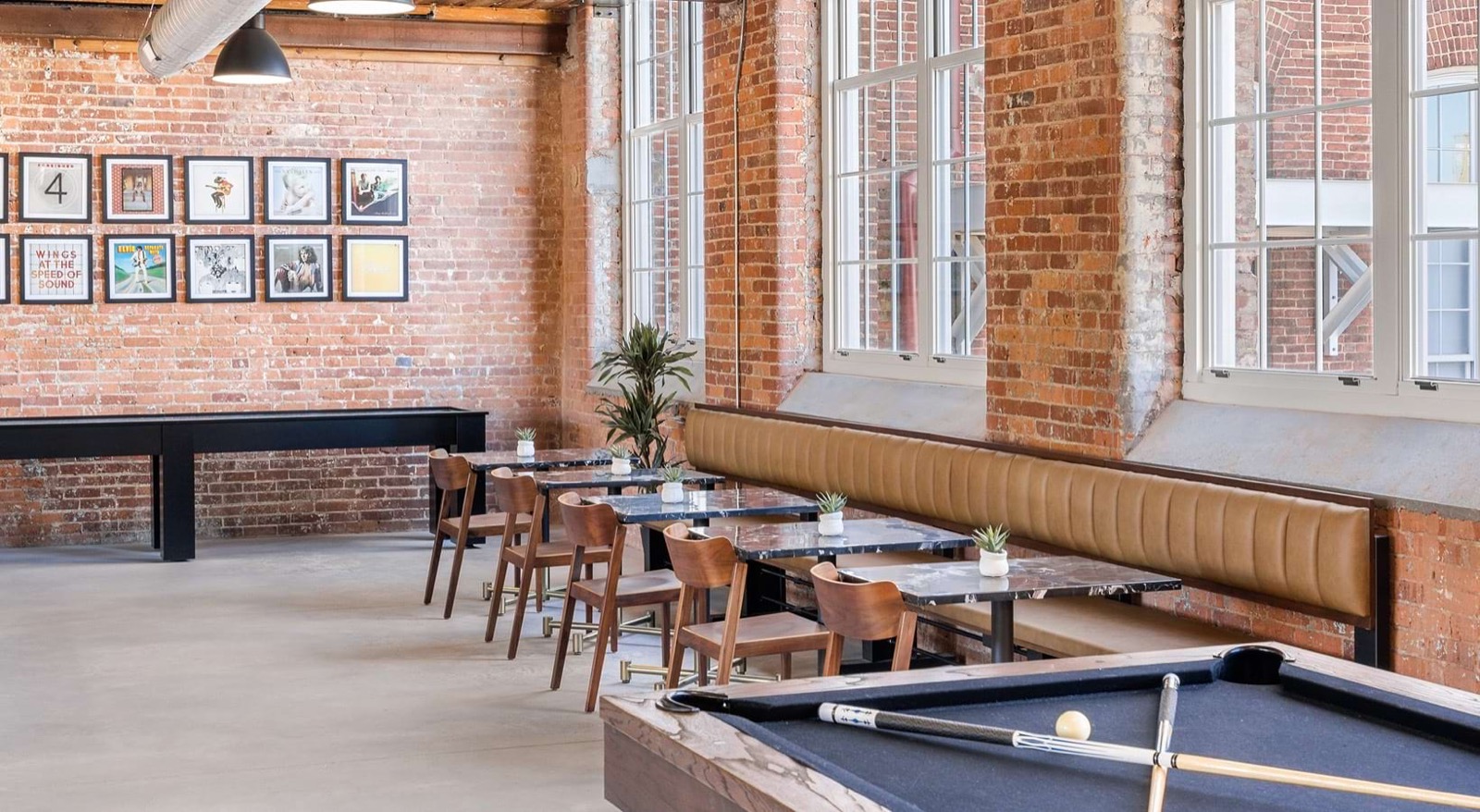 A game room with a pool table, leather banquette seating, marble caf&eacute; tables, and a wall of framed album covers beside tall brick-framed windows