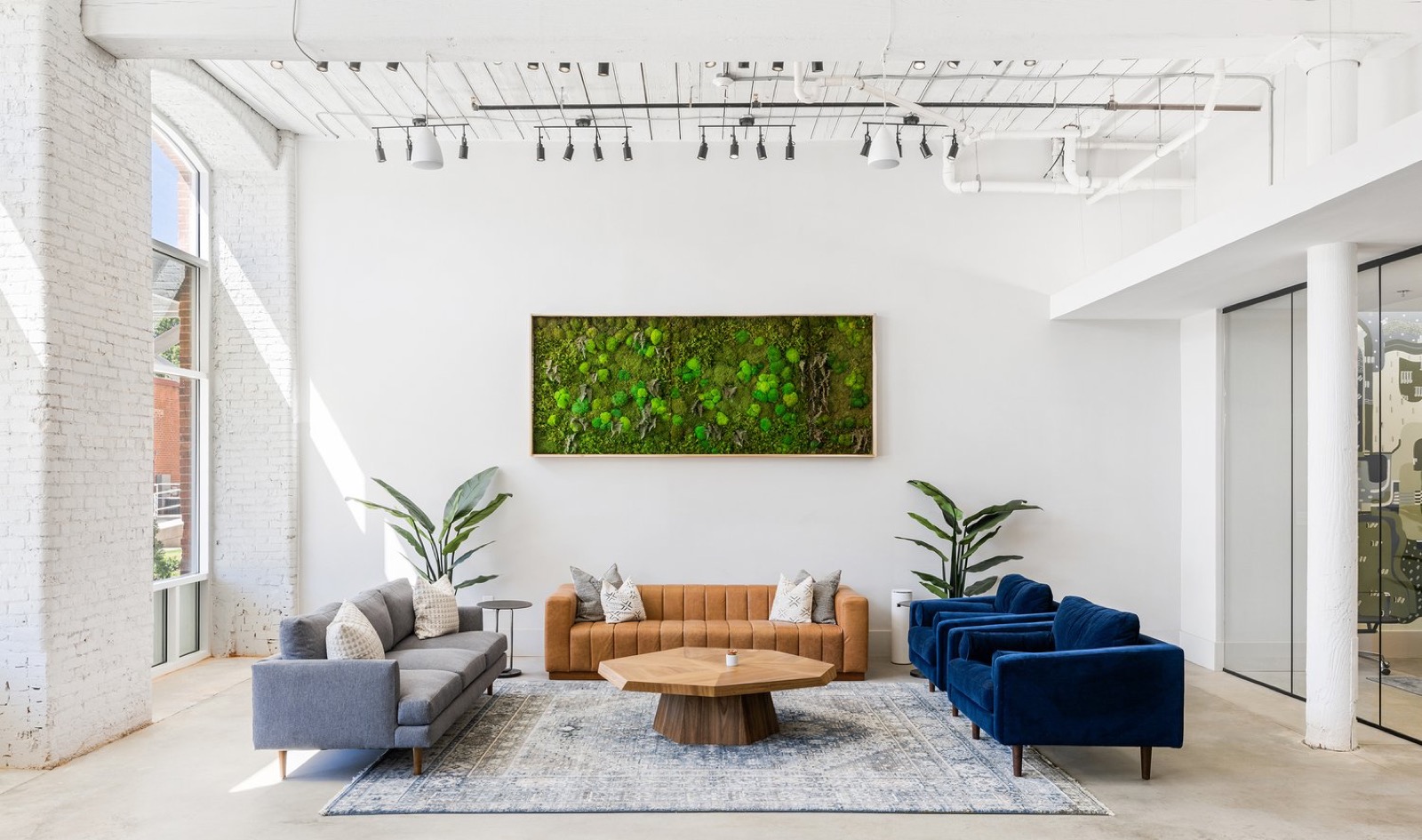 A bright lounge with a gray sofa, a tan leather sofa, blue velvet chairs, an octagonal coffee table, and a moss wall beneath white-painted beams
