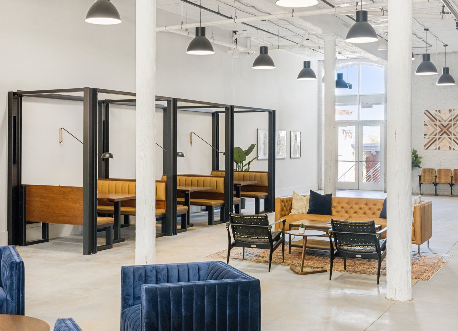 Steel-framed booths with tan leather seating beside a lounge of blue velvet chairs and a tan tufted sofa between white columns