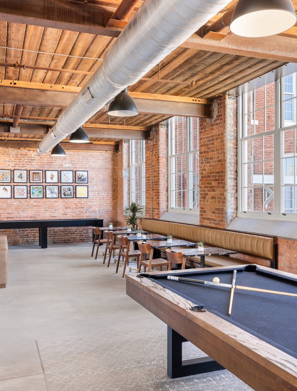 A pool table in the foreground of a game room with a wall of framed album covers, a banquette, and tall windows