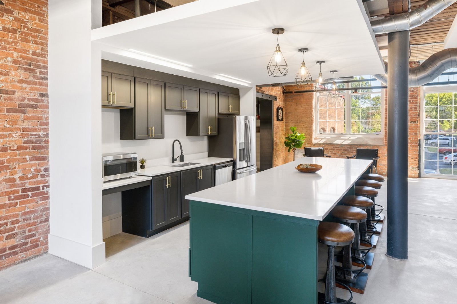 A kitchen with dark cabinets, brass pulls, and a long white-topped island in deep green with leather stools beside brick walls