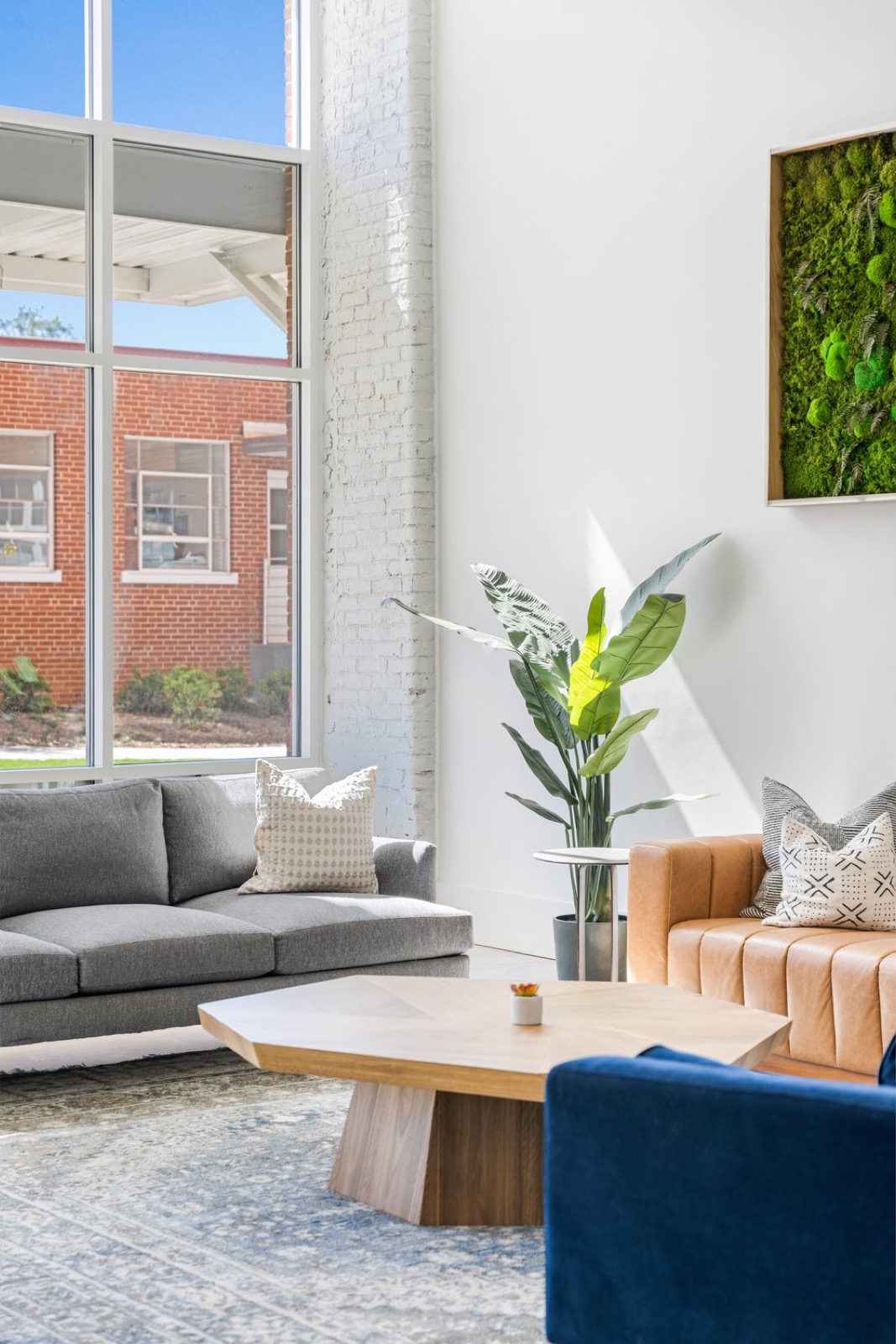 A gray sofa, a tan leather sofa, and an octagonal wood coffee table beneath a moss wall beside a tall window