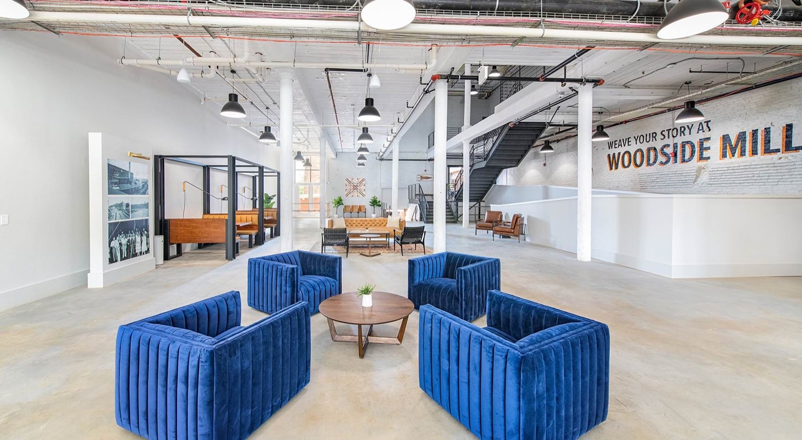 Four blue velvet chairs around a round table in the open lobby, with steel-framed booths, a staircase, and a mural reading Weave Your Story at Woodside Mill