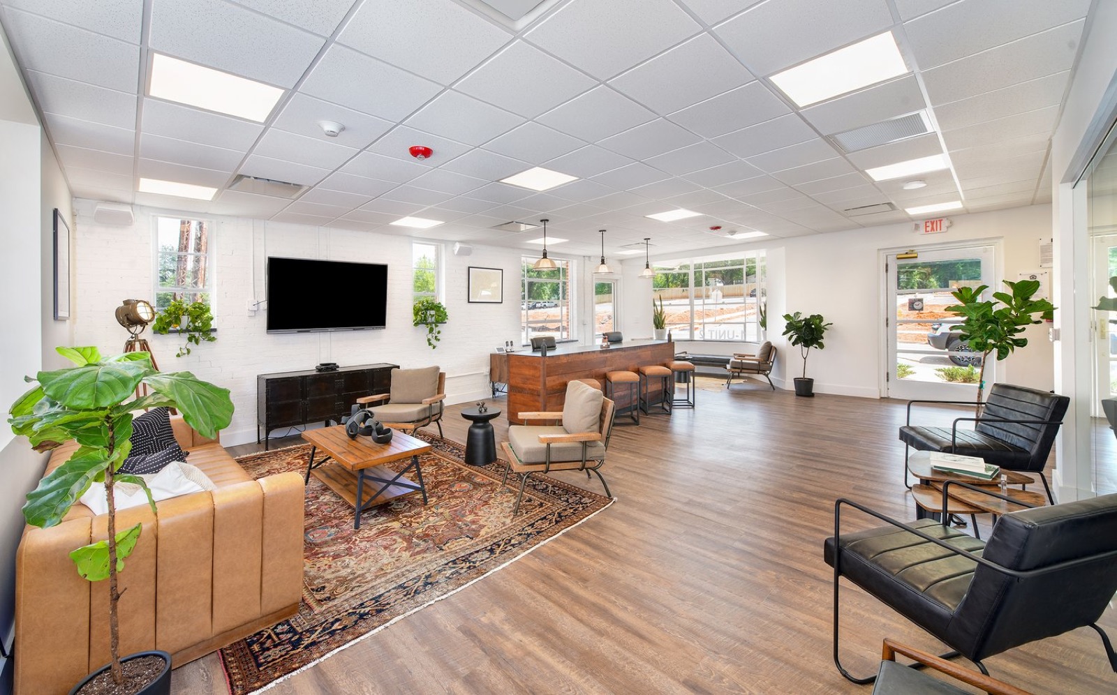 The leasing lounge with a tan sofa, a patterned rug, a reclaimed-wood reception counter, and black leather chairs beneath a ceiling of white tiles