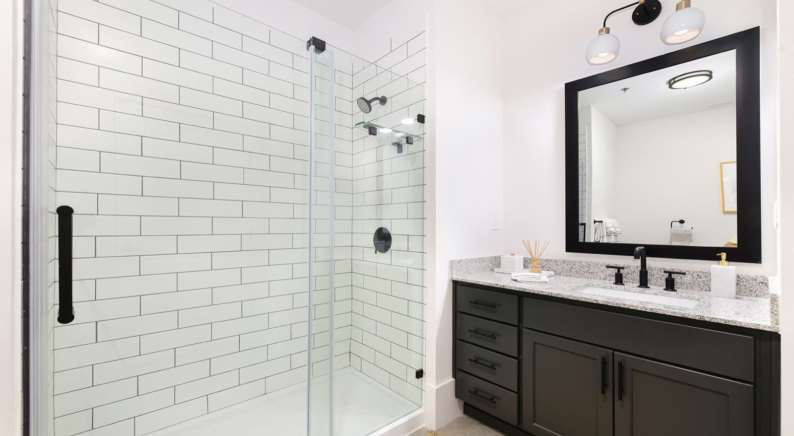 A bathroom with a glass-enclosed shower of white subway tile, a dark green vanity with a granite top, and a black-framed mirror