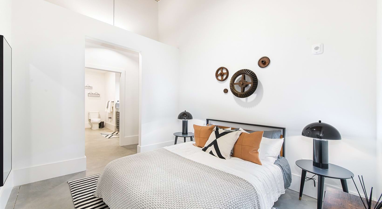 A bedroom with a gray-patterned bedspread, tan leather pillows, two dome lamps, and rusted mill gears mounted on the wall, with a bathroom beyond